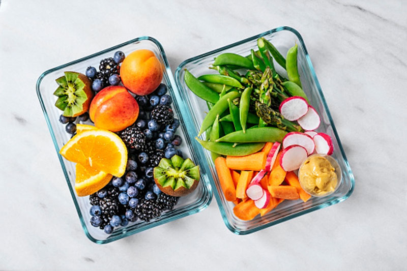 Two glass lunch containers: one with blueberries, blackberries, kiwi fruit and orange slices, the other with sliced up asparagus, snow peas, carrots and radishes